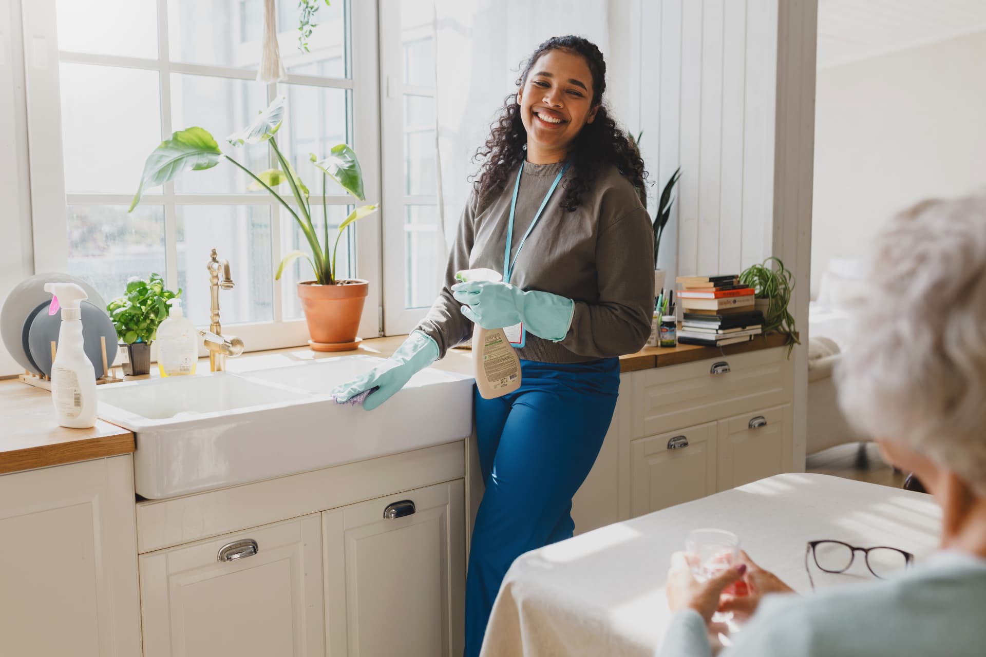 A smiling volunteer in rubber gloves cleaning a kitchen sink while an older neighbor sits nearby